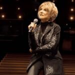 Jeannie Seely holds a microphone while seated on stage at the Grand Ole Opry, where a public memorial service will honor her 5,398th show.